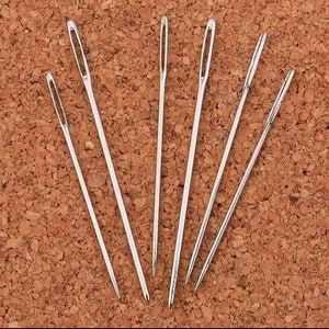 Może przedstawiać: Zestaw siedmiu srebrnych igieł do szycia, ułożonych wachlarzowo na brązowej powierzchni korkowej.