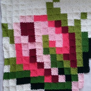 May include: A crocheted blanket with a pixelated design of a flower in shades of pink, red, green, and black.