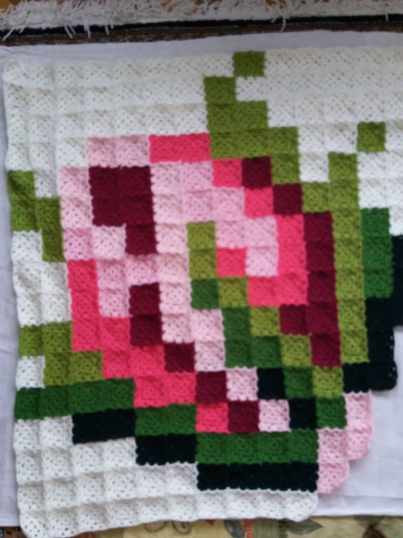 May include: A crocheted blanket with a pixelated design of a flower in shades of pink, red, green, and black.