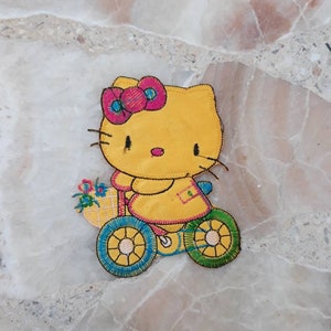 Könnte beinhalten: Gelber gestickter Bügelaufkleber mit Hello Kitty auf einem Dreirad mit einem Blumenkorb. Das Dreirad hat grüne Räder mit gelben Speichen und blauen Verzierungen.