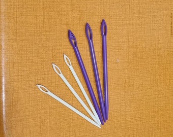 May include: Five plastic needles, four white and one purple, arranged in a fan shape on a brown surface.