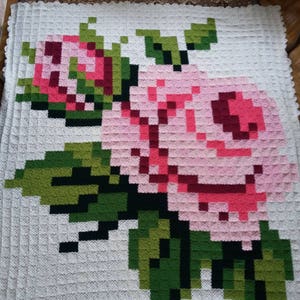 May include: A white crocheted blanket with a pink and green floral design. The flowers are in a pixelated style, resembling a video game.