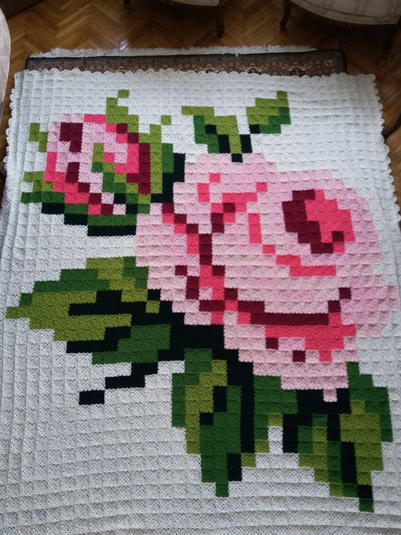 May include: A white crocheted blanket with a pink and green floral design. The flowers are in a pixelated style, resembling a video game.