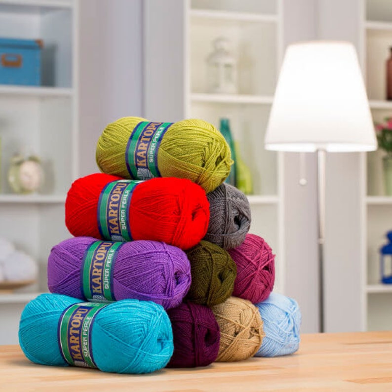 May include: A pyramid of colorful yarn skeins in shades of green, red, purple, and teal. Each skein is labeled with a blue and green band. The yarn is stacked on a wooden surface, with a lamp and shelves in the background.