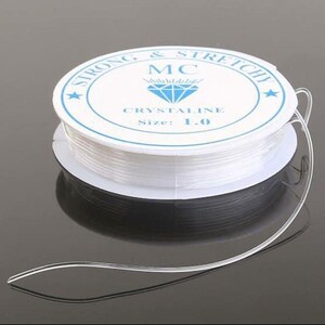 May include: A spool of clear, stretchy beading cord. The white label reads "STRONG & STRETCHY" and "CRYSTALINE Size: 1.0". The cord is wound around a white spool with a blue border and a diamond graphic. The cord is partially unwound.