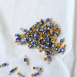 May include: A large group of gold safety pins with blue and white evil eye beads. The pins are scattered on a white fabric surface.