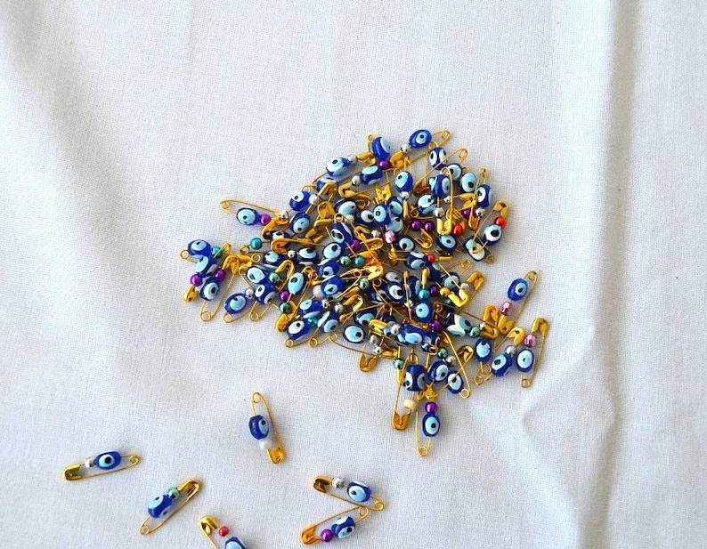 May include: A large group of gold safety pins with blue and white evil eye beads. The pins are scattered on a white fabric surface.