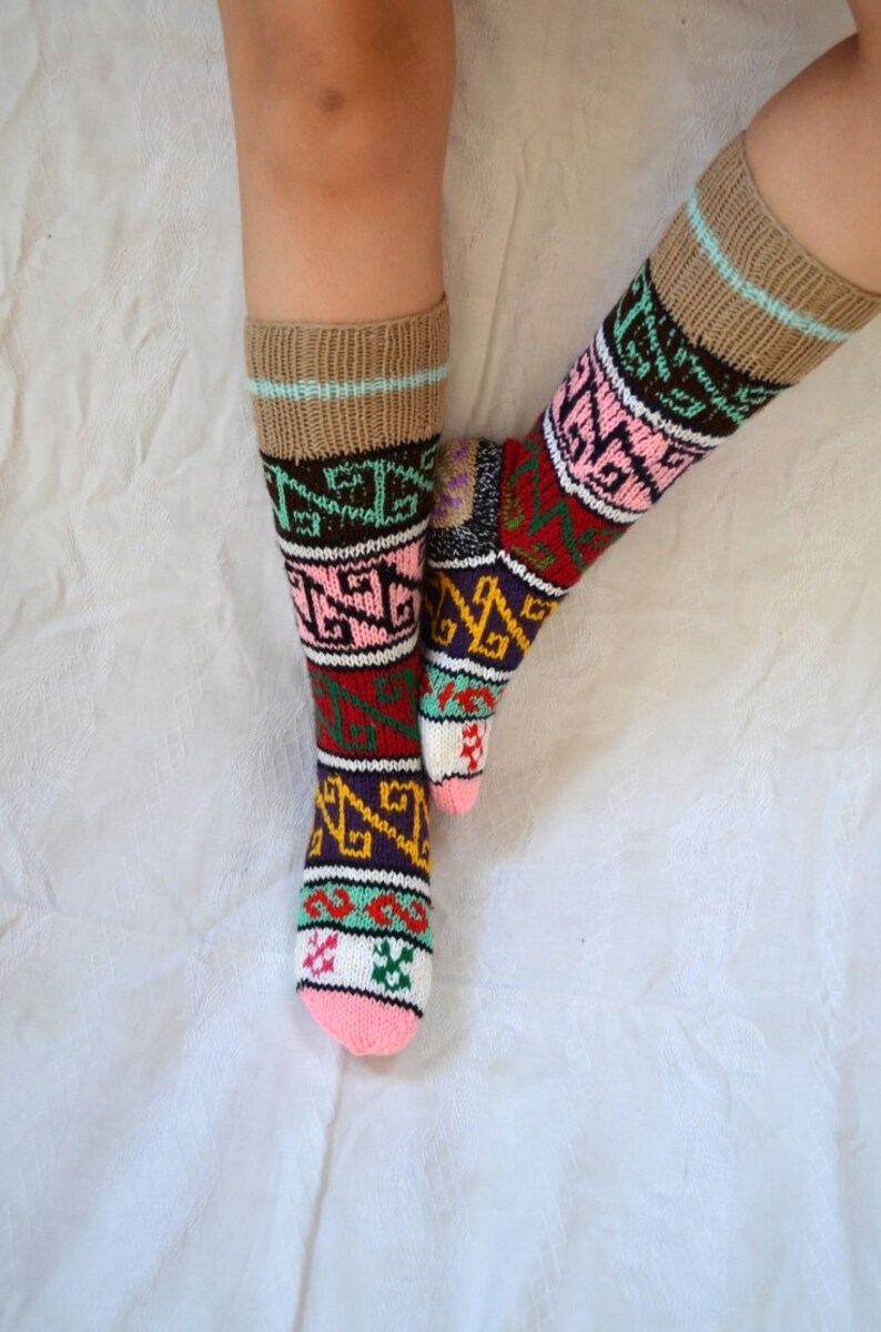 May include: A pair of colorful knitted socks with geometric patterns in shades of pink, green, red, yellow, and brown. The socks are worn on a person's legs.