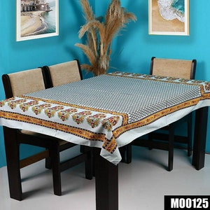 May include: A white tablecloth with a black and white geometric pattern and a yellow and orange floral border. The tablecloth is on a wooden table with four chairs. The table is in a room with blue walls and two framed pictures.