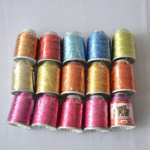 May include: A collection of 15 spools of metallic embroidery thread in various colors, including gold, silver, red, blue, and pink. The spools are wrapped in clear plastic and labeled with the brand name "Kaplan".