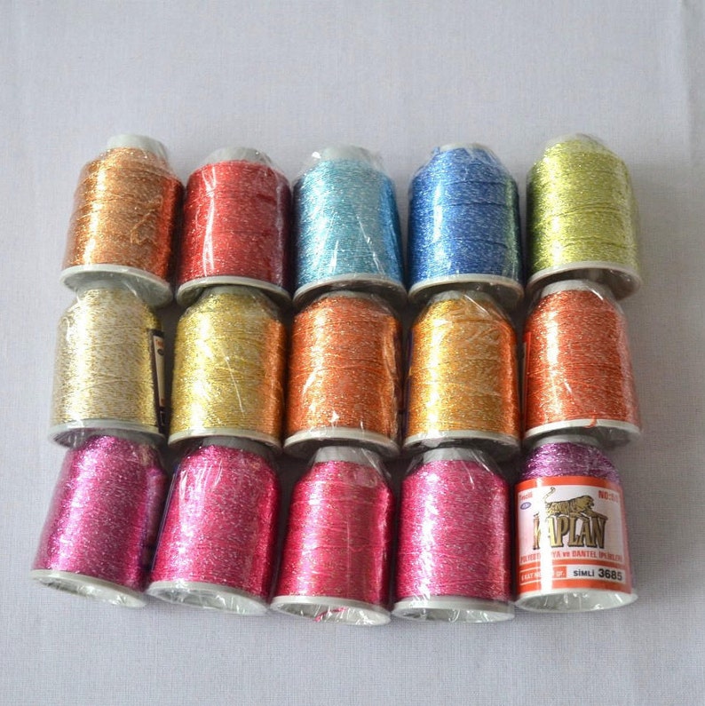 May include: A collection of 15 spools of metallic embroidery thread in various colors, including gold, silver, red, blue, and pink. The spools are wrapped in clear plastic and labeled with the brand name "Kaplan".