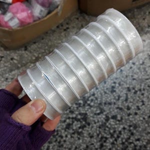 May include: A stack of clear, plastic ribbon spools, tightly wound and packaged. The spools are held in a hand, with other craft supplies visible in the background. The ribbon appears to be for crafting or gift wrapping.