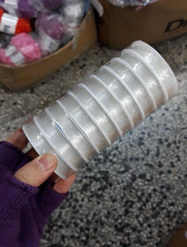 May include: A stack of clear, plastic ribbon spools, tightly wound and packaged. The spools are held in a hand, with other craft supplies visible in the background. The ribbon appears to be for crafting or gift wrapping.