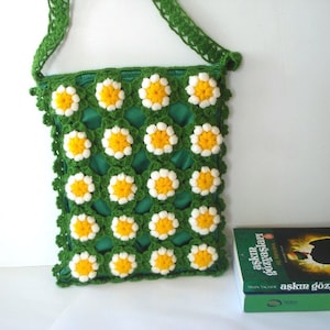 May include: A green crochet shoulder bag with a daisy pattern. The bag features a green strap and a green interior. The daisy pattern has white petals and yellow centers. A book with green cover and Turkish text is next to the bag.