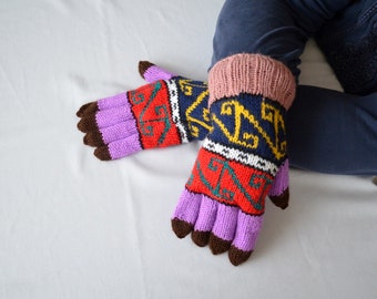 May include: A pair of knitted gloves with a colorful geometric pattern. The gloves are purple, red, blue, yellow, white, and brown. The gloves are worn by a person sitting on a white surface.