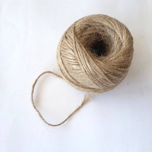 May include: A ball of natural twine or string, made from jute or hemp, is shown on a white background. The twine is brown and has a rough texture. A small length of twine is unraveled from the ball.