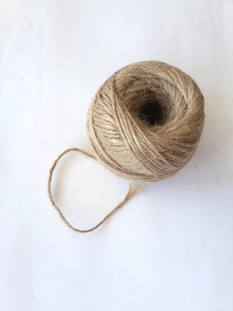 May include: A ball of natural twine or string, made from jute or hemp, is shown on a white background. The twine is brown and has a rough texture. A small length of twine is unraveled from the ball.