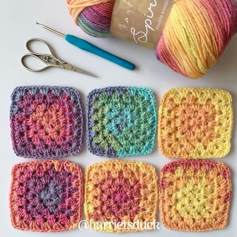 May include: Six colorful crochet granny squares in various shades of pink, purple, orange, yellow, green, and blue. The yarn is a gradient color and the squares are arranged in two rows of three. The yarn ball is labeled "Spirit double knit" and is in the background.