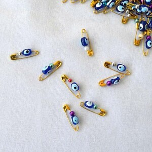 May include: A collection of gold safety pins with blue and white evil eye charms. The charms are attached to the pins with a small silver bead.