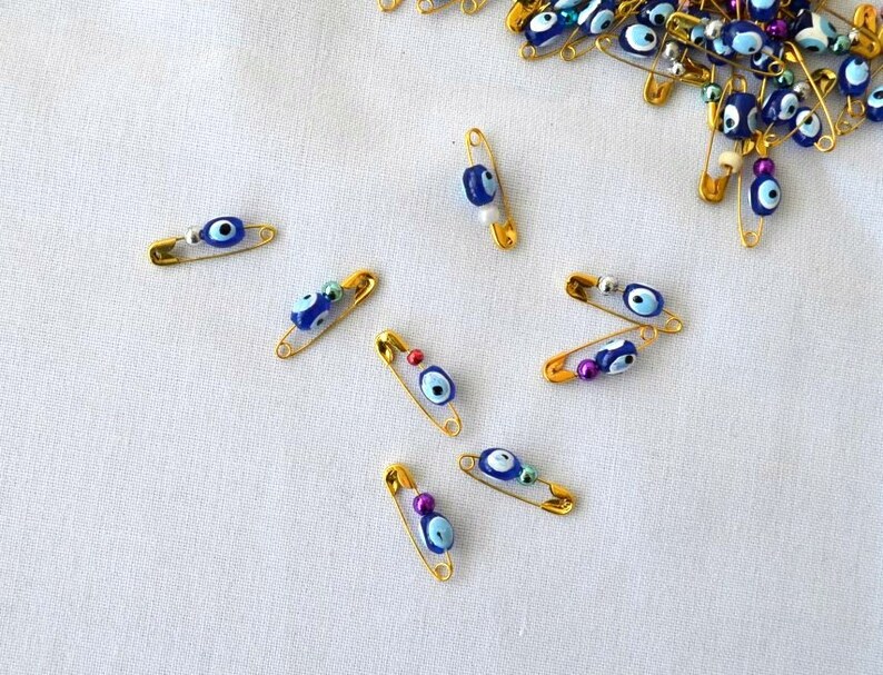 May include: A collection of gold safety pins with blue and white evil eye charms. The charms are attached to the pins with a small silver bead.