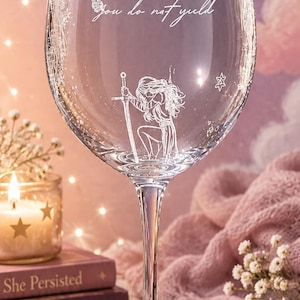 May include: A clear glass wine goblet featuring white etched designs of stars, a sun, and a figure kneeling with a sword. The text "You do not yield" is etched in a cursive script across the center of the glass bowl above the figure.