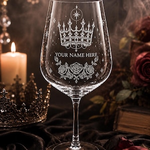 May include: A clear glass wine glass featuring an etched design of a crown, stars, and three roses. The text "YOUR NAME HERE" is engraved in the center of the glass. The stem and base are transparent, and the bowl has a tapered, rounded shape.