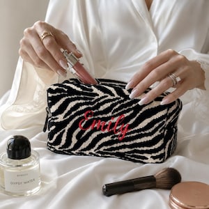 May include: A rectangular cosmetic bag featuring a black and white zebra print pattern with the name Emily embroidered in red script on the front. The bag has a black zipper closure and is shown being filled with a small tube of lip gloss.