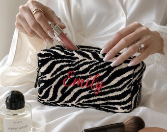 May include: A rectangular cosmetic bag featuring a black and white zebra print pattern with the name Emily embroidered in red script on the front. The bag has a black zipper closure and is shown being filled with a small tube of lip gloss.