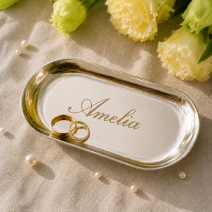 Könnte beinhalten: Ein ovales Tablett aus silberfarbenem Metall mit dem eingravierten Namen Amelia in Schreibschrift. Zwei goldfarbene Ringe, einer schlicht und einer mit kleinen Steinen besetzt, liegen auf der Oberfläche. Das Tablett ist von gelben Blumen und Perlen umgeben.