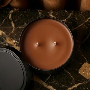 Peut inclure: Une boîte à bougie ronde en métal noir contenant de la cire brune lisse avec deux mèches en coton blanc. Le couvercle est placé à gauche du récipient ouvert, et un petit emblème carré doré avec un blason en relief est situé à droite.