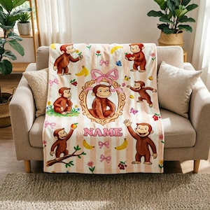 Könnte beinhalten: Eine weiche Wohndecke mit einem wiederkehrenden Muster aus braunen Affen, gelben Bananen, rosa Schleifen und bunten Blumen auf einem hellrosa und weiß gestreiften Hintergrund. Ein goldener Rahmen zeigt einen Affen mit dem Text NAME darunter.