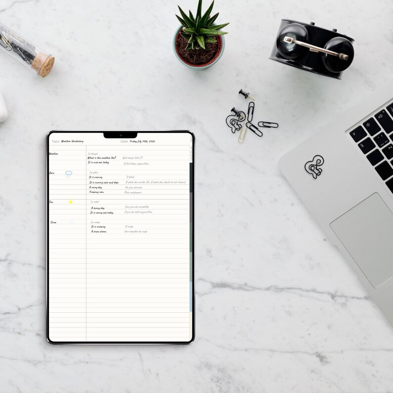 May include: A digital tablet displaying a weather vocabulary lesson plan with lined paper, a small potted succulent, a black alarm clock, paper clips, and a laptop with a sticker. The tablet screen shows text including Weather Vocabulary.