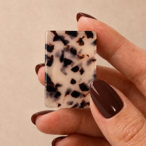 May include: A rectangular acrylic piece featuring a cream-colored base with an irregular pattern of dark brown and black tortoiseshell-style spots. The object has smooth, rounded corners and a flat, polished surface with a glossy finish.