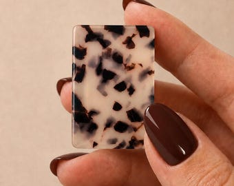Peut inclure: Une pièce rectangulaire en acrylique présentant une base de couleur crème avec un motif irrégulier de taches façon écaille de tortue marron foncé et noir. L'objet a des coins arrondis et une surface plate polie avec une finition brillante.
