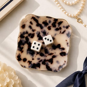 May include: A square, wavy-edged trinket tray featuring a cream and dark brown tortoiseshell pattern with a thin gold-toned rim. Two white six-sided dice with black pips are resting in the center of the tray, displaying the numbers three and four.