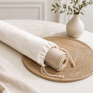 May include: A cylindrical off-white fabric drawstring bag containing a tightly rolled beige mat. The bag features a simple cinched top with matching fabric cords, partially revealing the rolled beige material tucked inside the protective sleeve.