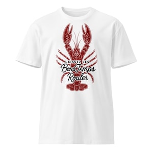 Puede incluir: Una camiseta blanca de manga corta con un gráfico detallado de una langosta de color rojo oscuro centrado en el pecho. Sobre la langosta aparece el texto negro "LAISSEZ LES BonsTemps Rouler" en una mezcla de fuentes de bloque y cursiva.