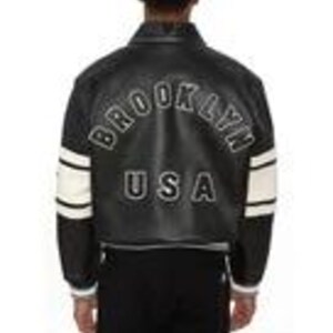 May include: A black varsity-style jacket featuring white leather sleeves with two horizontal black stripes. The back displays the text BROOKLYN in a white arched font above the letters USA in a bold white font, all set against a black leather body.