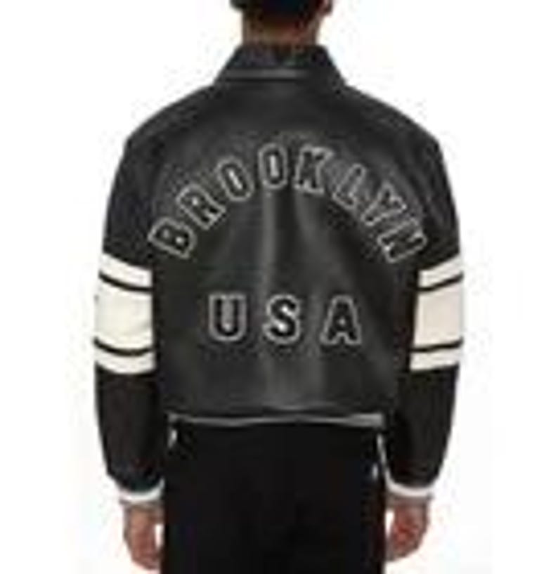 May include: A black varsity-style jacket featuring white leather sleeves with two horizontal black stripes. The back displays the text BROOKLYN in a white arched font above the letters USA in a bold white font, all set against a black leather body.