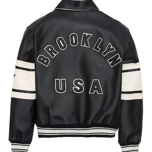 May include: A black varsity-style jacket featuring white stripes on the sleeves and a ribbed hem with white stripes. The back displays the text BROOKLYN in an arched font and USA below it, both in white lettering with black outlines on the black leather.