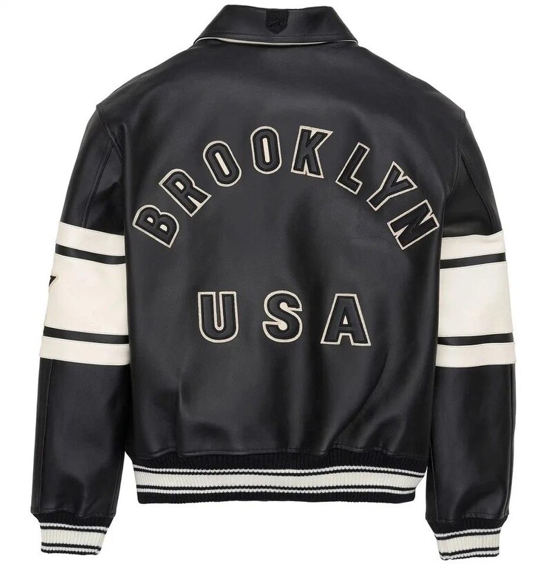 May include: A black varsity-style jacket featuring white stripes on the sleeves and a ribbed hem with white stripes. The back displays the text BROOKLYN in an arched font and USA below it, both in white lettering with black outlines on the black leather.