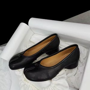 Könnte beinhalten: Ein Paar schwarze Lederschuhe mit abgerundeter Spitze, einem geschlitzten Vorderteil und einem niedrigen Blockabsatz. Das Innere der Schuhe ist in einer beigen Farbe gefüttert. Die Schuhe haben eine glatte Lederoberfläche und ein einfaches Slip-on-Design.