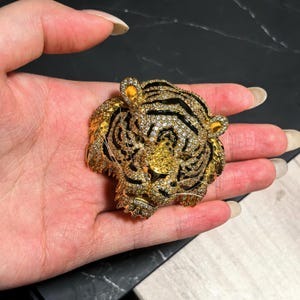 May include: A gold-toned tiger head brooch featuring a dense pav&eacute; of clear, sparkling crystals and black enamel stripes. The tiger's face is detailed with a yellow crystal nose and pointed ears, creating a textured, three-dimensional appearance.