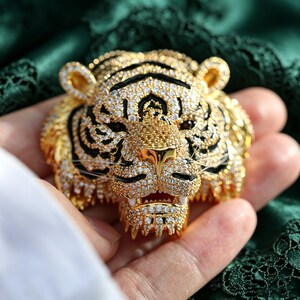 May include: A gold-toned tiger head brooch featuring intricate detailing with clear pav&eacute;-set crystals and black enamel accents. The design captures the feline's facial features, including pointed ears, a textured snout, and visible white fangs.