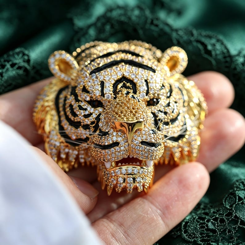 May include: A gold-toned tiger head brooch featuring intricate detailing with clear pav&eacute;-set crystals and black enamel accents. The design captures the feline's facial features, including pointed ears, a textured snout, and visible white fangs.