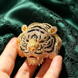May include: A gold-toned tiger head brooch featuring intricate pav&eacute;-set clear crystals and bold black enamel stripes. The tiger has a detailed face with visible whiskers, pointed ears, and an open mouth showing teeth, crafted with a metallic finish.