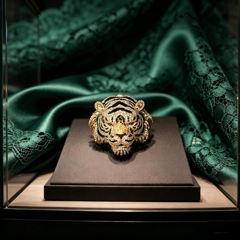 May include: A gold-toned tiger head brooch or ornament featuring intricate black enamel detailing and encrusted with numerous clear, sparkling crystals. The tiger head is rendered in a realistic style with pointed ears and a snarling expression.
