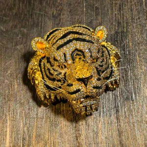 May include: A tiger head brooch crafted in gold-tone metal, fully encrusted with clear sparkling rhinestones. The design features black enamel stripes across the forehead and cheeks, with bright yellow accents inside the ears and eyes.