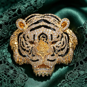 May include: A gold-toned tiger head brooch featuring a dense pav&eacute; of clear white crystals and black enamel stripes. The tiger has amber-colored eyes, a textured golden nose, and visible white fangs, creating a detailed and ornate accessory.