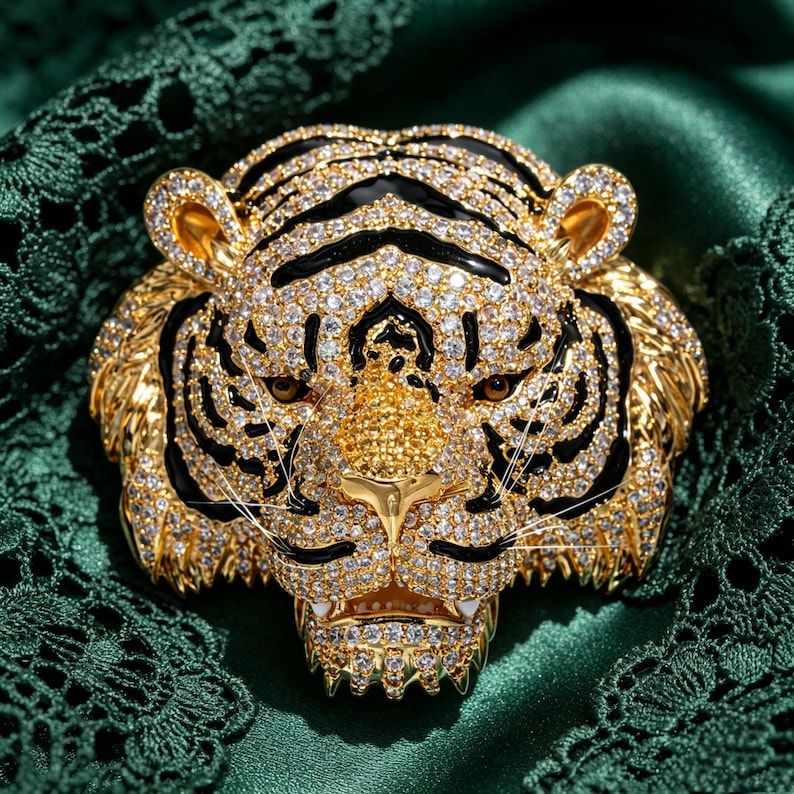 May include: A gold-toned tiger head brooch featuring a dense pav&eacute; of clear white crystals and black enamel stripes. The tiger has amber-colored eyes, a textured golden nose, and visible white fangs, creating a detailed and ornate accessory.